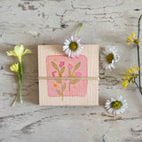 Eine Draufsicht auf ein kleines, rosagrün mit Blüten bemaltes Holzquadrat. Runherum liegen Gänseblümchenköpfe und Primeln.