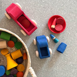 Verschiedene Pikler Spielmaterialien aus Holz in den Farben rot und blau stehen am Boden, daneben ein Korb mit weiterem bunt lasiertem Holzspielzeug darin.