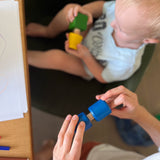Ein Kleinkind versucht eine Holzschraube und eine Holzmutter von Nic Toys ineinander zu schrauben, daneben schrauben erwachsene Hände das blaue Schrauben-Mutter Set zusammen.