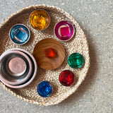 7 Lichtbauwürfel von Bauspiel in verschiedenen Farben liegen in kleinen Klangschalen als Edelstahl oder einem Holzteller, die wiederum in einem Korb aus Naturmaterial stehen.