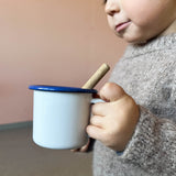 Ein Kind hält eine weiße Kindertasse aus Emaille mit blauem Rand.