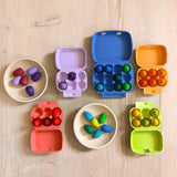Kleine bunte Holzeier in 12 Farben von Grapat aufgeteilt in bunten, farblich passenden Eierkartons. Zwei runde Naturholzschalen stehen dazwischen, ebenfalls mit bunten Mini-Holzeiern gefüllt.