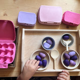 Auf einem Holztablett sind Holzschalen mit kleinen lila Holzeiern, dazwischen kleine Kochlöffel, rundherum stehen kleine Eierkartons in zartem lila, rosa und rot. Kinderhände, die nach einem Holzei greifen.