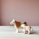 Ostheimer Spielfiguren aus Holz. Die Kuh mit braunen Flecken steht mit erhobenem Kopf und Blick nach links. Ein kleines Kalb steht seitlich mit Blick zur Kuh daneben.
