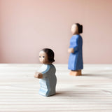 Die Ostheimer Spielfigur Wickelkind steht auf einer Holzplatte und schaut nach links. Im Hintergrund rechts ist unscharf die Ostheimer Holzfigur Mutter zu sehen.