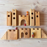 Alle zehn Teile der Ritterburg Holzbausteine von Bauspiel aufgelegt auf dem Boden, als Größenvergleich liegen verschiedene Grapat und Playmobil Figuren darauf.