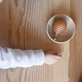 Der Arm eines Kindes streckt sich nach einer runden, flachen Holzschale, in der die Rassel Kugelrund liegt.