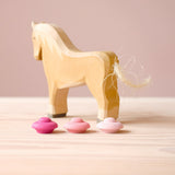 Eine Holzpferd Spielfigur, davor drei Mandala Steine von Grapat "Flower" - also drei kleine Blüten aus Holz in verschiedenen Rosatönen.