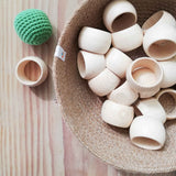 Serviettenringe aus Holz, ganz naturblessen liegen in einem orb, daneben liegt ein Serviettenring und ein gräner Häkelball von Kokomoo auf dem Tisch.