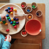 Grapat "Bowls and Acorns" Sortierspiel: das sind kleine Holzeicheln in Regenbogenfarben und 6 bunte Holzschälchen - hier liegen sie auf einem kleinen Tisch, dazu eine Holzschale und ein oranges Jutekörbchen.