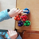 Ein Kind sortiert die bunten kleinen Holzeicheln in Regenbogenfarben vom Sortierspiel Grapat "Acorns & Bowls". Es legt grad die letzte blaue Eichel mit der Holzzange auf das blaue Schälchen.