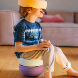 Ein Kleinkind sitzt auf einem Balancierhocker aus zwei pastellfarbenen Stapelsteinen, auf seinem Kopf trägt es den pastellfarbenen  Stapelstein® in orange.
