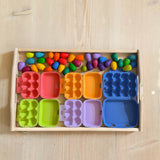 Auf einem Holztablett stehen 5 bunte Mini-Eierkartons. Daneben liegen 36 Mandalaeier von Grapat in Regenbogenfarben.