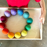 Ein Kind trägt ein Holztablett, darauf sind 12 Schälchen aus Holz von Grapat, gefüllt mit 12 Filzbällen von Kokomoo in Regenbogen Farben, aufgestellt im Kreis.