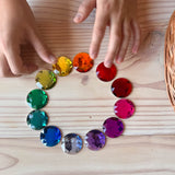 Die zwölf Maxi Funkelsteine von Bauspiel farblich sortiert im Kreis aufgelegt. Kinderhände greifen danach.