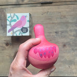 Der kleine Kreisel aus Holz Chill Pink Bird von Grapat wird in der Hand gehalten, man sieht die Rückseite des Vogels, liebevoll bemalt. Am Tisch darunter liegt die Verpackung.