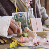 Eine Kinderhand hält eine kleine Holzfigur und stellt sie auf einer Tischfläche ab. davor steht ein Ostheimer Lämmchen mit rosa Halsband und es liegen Blüten und Grapat Holztulpenköpfe auf der Tischdecke.
