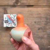Blick von oben, eine Hand hält den Kreisel Mellow Orange Bird von Grapat. Wir sehen die orange, grünlich, rötliche Bemalung vom Vogel-Kreisel aus Holz.Darunter am Tisch liegt die Verpackung.
