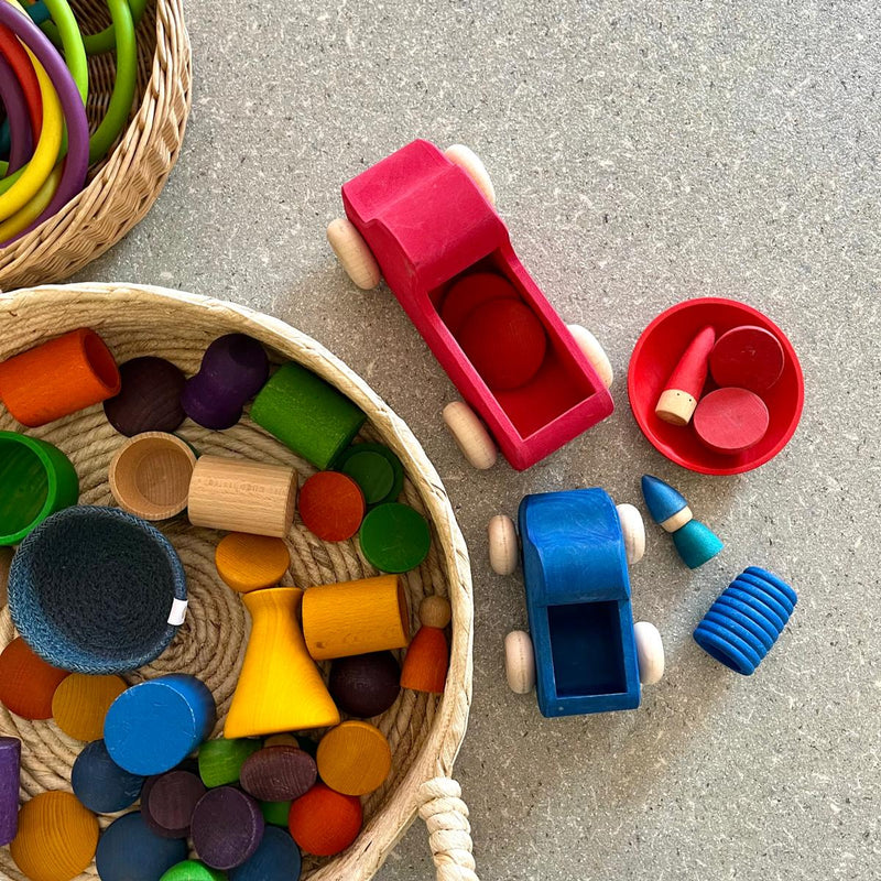 Ein großer Tablettkorb mit Tragegriffen gefüllt mit Pikler Spielmaterialien von Kokomoo, daneben Grimms erste Lastwägen in blau und rot.