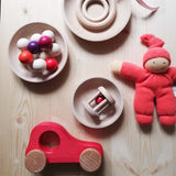 Babyspielsachen aus Holz von Kokomoo verteilt in Holzschalen, ein Kugelgreifling, ein Holzauto, eine kleine Rassel, daneben ein Wichtel von Nanchen in Rot.