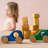Ein blaues und ein grünes Baby-Holzauto zum Greifen von Kokomoo, dahinter Holzbausteine und ein Baby.