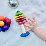 Ein kleine Klangschale, die Regenbogenrassel Bäumchen von Nic Toys und der Kugelgreifling von Kokomoo auf einer weißen Babydecke verstreut. Eine Babyhand greift nach der Rassel.