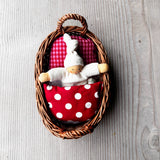 Das Wichtelnest von Kokomoo in rot: ein Weidenkörbchen als Puppenbett, in dem ein weißer Nanchen Wichtel in einer rot gemusterten Bettwäsche kuschelt. 