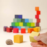 Zwei gelbe Würfel des Grimms Bausteine Set "Regenbogen Mosaik" ganz von nahem zu sehen, daneben ein Schneckenhaus als Größenvergleich, im Hintergrund unscharf die restliche Bauklötze bunt zusammenebaut.