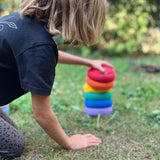 Ein Kind baut einen farblich sortierten Turm aus sechs Stapelsteinen Mini in Regenbogenfarben.
