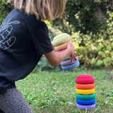 Ein Kind hält einen farblich sortierten Turm aus sechs Stapelsteinen Mini in Pastell, im Begriff diesen auf einen Turm  in Regenbogenfarben zu setzen.