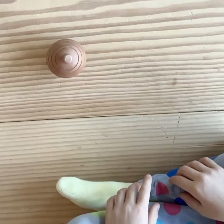 Die Rassel Kugelrund, gesehen von oben, kreiselt vor einem Kind im Kreis herum. Dann Zeitlupe. Das Kind streckt den Arm nach der Rassel aus, hält diese aber nicht an, sondern bleibt in der Beobachtung.