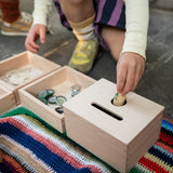 Ein Kind spielt mit der Grapat "Permanence Box" und versucht, ob eine kleine Spielfigur aus Holz durch das Loch passt. Daneben sind andere Materialien zum Versuchen angeboten.