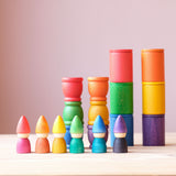 Die Grapat "Rainbow Tomtens", kleine Holzfiguren zum Spielen in Form von Zwergen in den sechs Regenbogenfarben nebeneinander aufgestellt, dahinter Grapat "Coloured Cups with Lid" Holzbecher und Grapat "Mates" Holztöpfchen in den gleichen Farben.