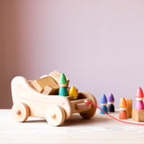Zwei Grapat Holzzwerge in Grün und Gelb sitzen in einem Nachziehwagen aus Holz, hinten ihnen Holzbausteine geladen, daneben stehen die erestlichen vier Zwerge, sodass alle Regenbogenfarben beisammen sind.