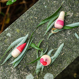 Die drei Holzfiguren "Familie Yay" von Grapat liegen gemeinsam mit Weidenblättern auf einem bemoosten Beton.