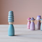 Vorne steht eine Grapat Fancy Nins Spielfigur in Blau mit einem farblich abgestimmten Hut, im Hintergrund weitere Spielfiguren aus Holz in Blau, Lila und Rosa.