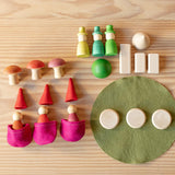 Das Spielset Freunde im Wald. Auf einem grünen Filzkreis liegen geometrische Formen - Kegel, Quader, Scheiben in Naturholz und eine grüne Halbkugel. Daneben stehen 3 handbemalte Holzpilze, drei grüne und 2 rote Holzkegel und es liegen 3 kleine pinkfarbene Filztaschen davor.