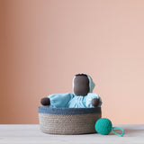 Die blaue Kuschelpuppe "Aqua" von Grimms aus Stoff sitzt in einem Korb mit blauem Rand. Daneben liegt ein türkiser Häkelball mit Schlaufe von Kokomoo.