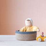 Die gelbe Kuschelpuppe "Sonne" von Grimms aus Stoff sitzt in einem Korb mit blauem Rand. Daneben steht ein kleines, gelbes Holzauto.