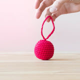 Eine Hand hält den Häkelball mit Schlaufe in der Farbe Kirsche an der Schlaufe hoch.