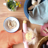Eine Kuschelpuppe liegt unter einem korallefarbenen, kleinen Stricktuch zugedeckt, ein weißer Nanchen Wichtel in seinem Stapelstein Bett liegt unter einem hellblauen Stricktuch.