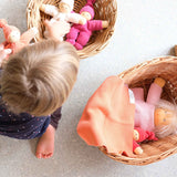 Ein Kind am Boden sitzend, zwei Körbe mit Puppen und Wichtel, auf einem Korb liegt auch ein kleines Stricktuch als Puppendecke.