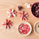 Blumen aus Mandala Legematerial aus Holz gelegt, in einer Holzschale daneben liegen "Flower Petals" Mandalasteine von Grapat, in einem pinken Jutekorb liegen "Pineapple" Mandalas.