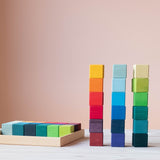 Grimms 36-tlg. Bausteine Set "Regenbogen Mosaik" teilweise im mitgelieferten Tablett, daneben 3 Türme aufgebaut zu je sechs Bauklötzen.