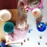 Zwei Kinder zu sehen von oben, die mit vielen Häkelbällen und Holzmadalas, verteilt in Körben, Kübel und Schalen am Boden spielen.