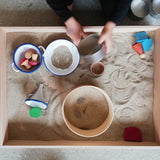 Blick von oben in eine bespielte Sandwanne mit Emaille Geschirr, Holzsieb, Holzfiguren, Blechdosen und weiteren sensorischen Spielmaterialien.