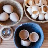 Drei Grapat Bowls Holzschälchen in einem blauen Jutekorb, daneben Körbchen mit Holzeiern und Serviettenringen - vorbereitete Spielsituation im Pikler Spielraum.