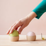 Eine Hand greift nach einem grünen Häkelball der in einem Grapat Holzschälchen liegt. Daneben steht ein Holzschälchen mit einem weißen Häkelball darin.