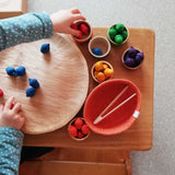 Ein Kind sortiert die bunten kleinen Holzeicheln in Regenbogenfarben vom Sortierspiel Grapat "Acorns & Bowls" farblich passend in die Holzschälchen.