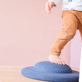 Ein Kleinkind steigt mit einem Fuß auf das Stapelstein® Balance Board in blau, das flach auf dem Boden liegt, mit dem Knubbel nach oben.